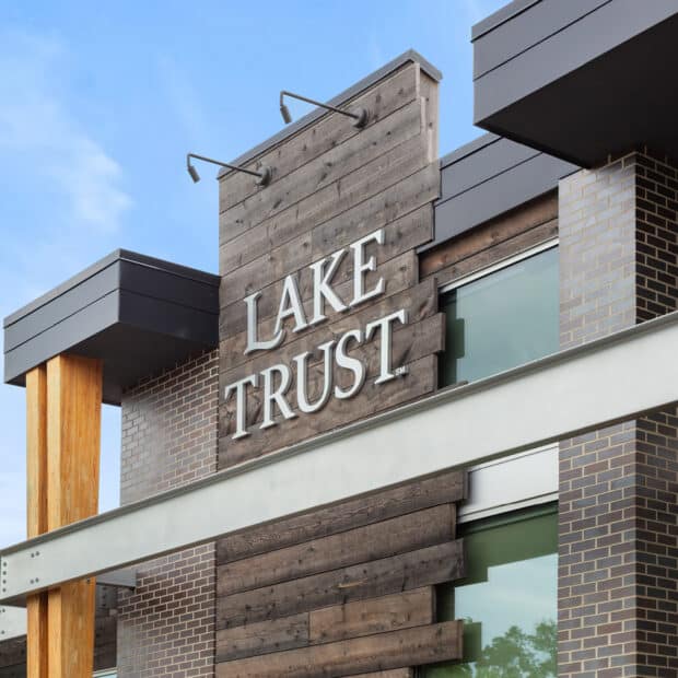 Modern building facade with "Lake Trust" sign on rustic wooden paneling, accented by glass windows and brickwork against a clear blue sky.