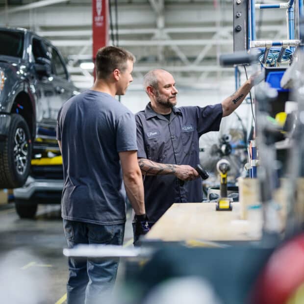 Two men stand in an auto repair shop. One points at equipment on a workbench, while a car is lifted in the background. The atmosphere is focused and collaborative.