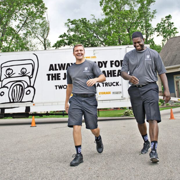 Two men in matching gray uniforms walk cheerfully in front of a large moving truck with a humorous illustration, conveying teamwork and friendliness.