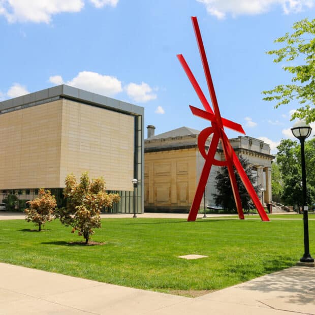 A vibrant red abstract sculpture stands prominently on a grassy lawn with modern and classical buildings in the background under a clear blue sky.